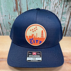 Op de afbeelding: Een donkerblauwe truckercap met een patch met een stier en de tekst "Bull City". De patch is oranje en wit met een gele achtergrond. De pet heeft een zwart mesh achterkant en een verstelbare band met het Richardson-logo en de tekst "Original 112 Trucker Adjustable".