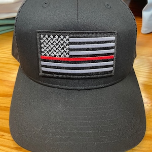 May include: Black mesh trucker hat with a black and white American flag patch with a red stripe across the middle.