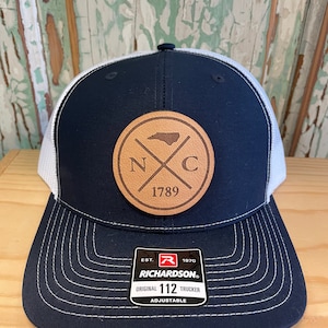 Może przedstawiać: Czapka trucker w kolorze granatowym i białym z brązową skórzaną naszywką przedstawiającą stan Karolina Północna i tekst "NC 1789". Czapka ma regulowany pasek z tekstem "Richardson. Original 112 Trucker Adjustable."