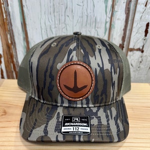May include: A camouflage trucker hat with a brown leather patch featuring a bird's foot design. The hat has a mesh back and a black label with the text "EST. 1970 RICHARDSON. ORIGINAL 112 TRUCKER ADJUSTABLE".