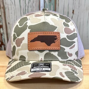 May include: A camouflage trucker hat with a brown leather patch featuring the outline of North Carolina. The hat has a mesh back and a curved brim. The patch is stitched onto the front of the hat. The hat has the text "RICHARDSON 112 TRUCKER" on a tag.