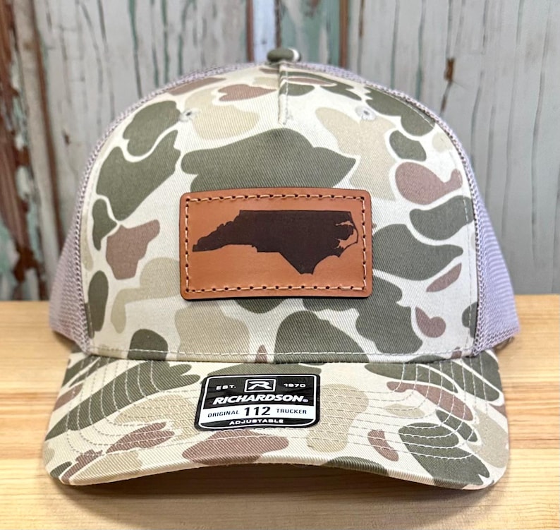 May include: A camouflage trucker hat with a brown leather patch featuring the outline of North Carolina. The hat has a mesh back and a curved brim. The patch is stitched onto the front of the hat. The hat has the text "RICHARDSON 112 TRUCKER" on a tag.