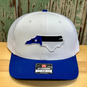 May include: White trucker hat with a blue and black North Carolina state outline patch. The hat has a royal blue brim and a mesh back. The hat has a Richardson 112 label on the side.