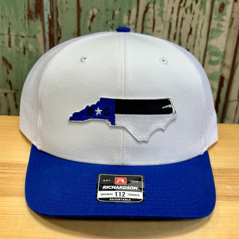 May include: White trucker hat with a blue and black North Carolina state outline patch. The hat has a royal blue brim and a mesh back. The hat has a Richardson 112 label on the side.