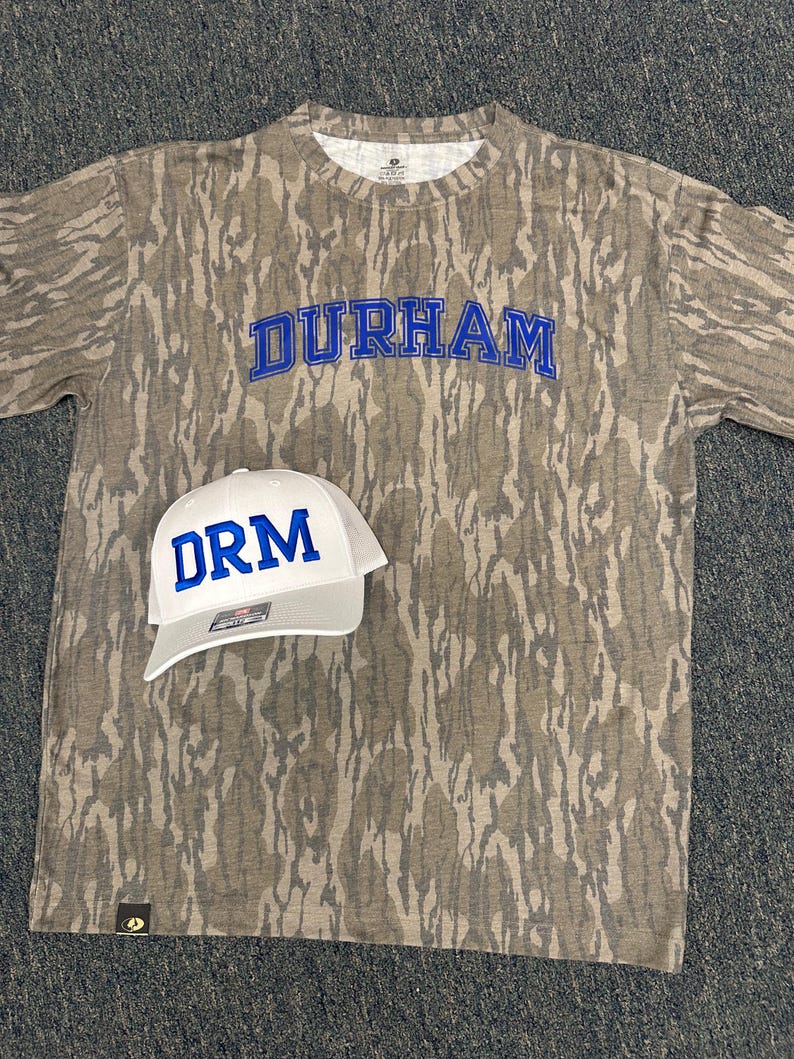 Op de afbeelding: Een camouflage-patroon T-shirt met het woord "DURHAM" in blauwe blokletters. Een witte baseballcap met "DRM" in blauwe blokletters ligt voor het shirt.