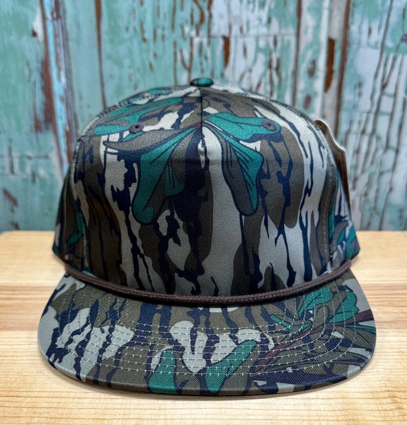 K&ouml;nnte beinhalten: Baseballkappe mit Tarnmuster und braunem Seildetail am Schirm. Die Kappe hat eine Mischung aus gr&uuml;nen, braunen und grauen Farben. Der Hut wird auf einer Holzoberfl&auml;che pr&auml;sentiert.
