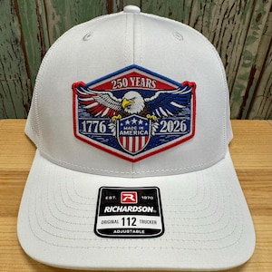 May include: White trucker hat with a mesh back and a curved brim. The front features a patch with an eagle, the text "250 YEARS", "1776", "MADE IN AMERICA", and "2026". A black and white tag reads "RICHARDSON ORIGINAL 112 TRUCKER ADJUSTABLE".