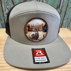 Puede incluir: Una gorra de béisbol de visera plana de color marrón claro y marrón con una parte trasera de malla negra. La gorra presenta un parche redondo con un ciervo de cola blanca en un entorno de bosque. El parche tiene un borde de cuero marrón. La gorra tiene una etiqueta roja y blanca que dice "Wear the Best, Richardson, Flat Bill Snapback, Est 1970."