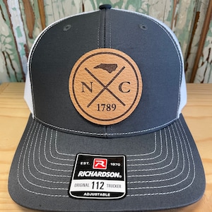 May include: A grey and white trucker hat with a leather patch showing the North Carolina outline, the letters "N" and "C", and the year "1789". The hat has a curved peak and an adjustable strap.