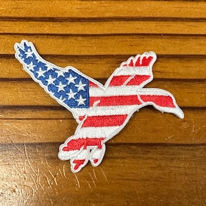 May include: An embroidered patch shaped like a flying duck, in red, white and blue. The duck is outlined in white and the American flag is depicted on its wings and body.