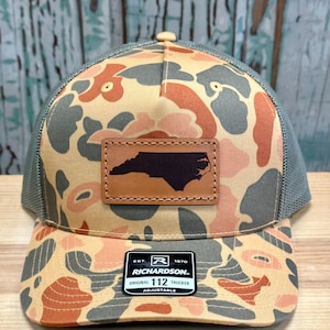 May include: A camouflage trucker hat with a brown leather patch featuring the outline of North Carolina. The hat has a tan, gray, and orange pattern, a gray mesh back, and a Richardson patch.