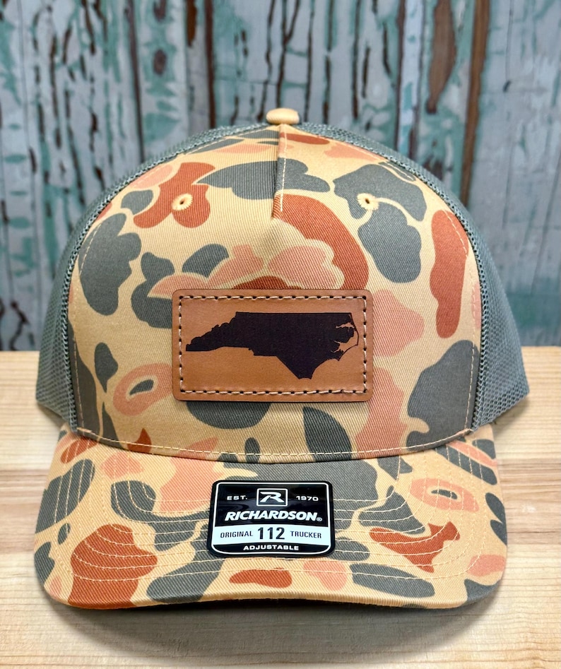 May include: A camouflage trucker hat with a brown leather patch featuring the outline of North Carolina. The hat has a tan, gray, and orange pattern, a gray mesh back, and a Richardson patch.
