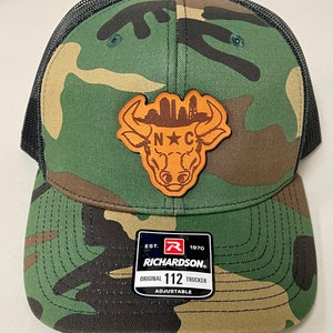 May include: Camouflage trucker hat with a brown leather patch featuring a bull silhouette and the letters "N C". The hat has a black mesh back and a curved brim. The Richardson brand tag is visible.