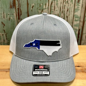 May include: Gray and white trucker hat with a North Carolina state flag patch. The hat has a black and white mesh back and a gray front with a black and white embroidered patch of the North Carolina state flag. The hat has a black adjustable strap with a black buckle and a white label that says "Richardson" and "Original 112 Trucker Adjustable".