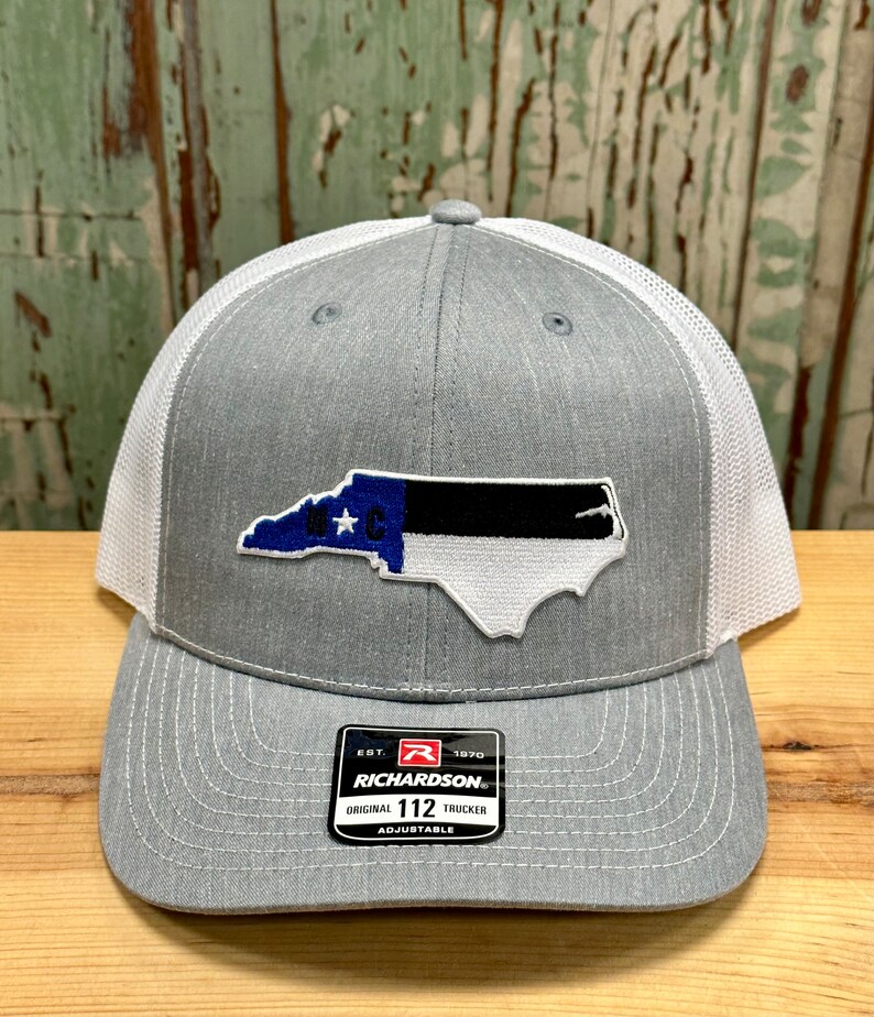 May include: Gray and white trucker hat with a North Carolina state flag patch. The hat has a black and white mesh back and a gray front with a black and white embroidered patch of the North Carolina state flag. The hat has a black adjustable strap with a black buckle and a white label that says "Richardson" and "Original 112 Trucker Adjustable".
