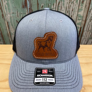 May include: A grey and black trucker hat with a brown leather patch featuring a bull silhouette and the text "Old Bull". The hat has a black mesh back and a label with the text "Richardson" and "Original 112 Trucker Adjustable".