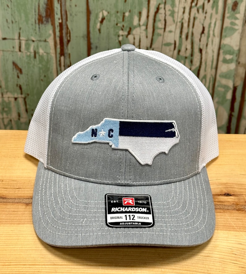 May include: A gray and white trucker hat with a blue and white North Carolina state flag patch. The hat has an adjustable strap and a Richardson 112 label.