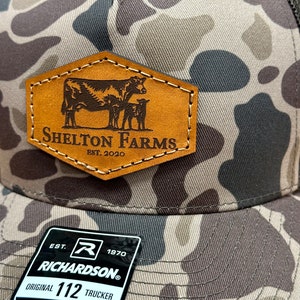 May include: A camouflage trucker hat with a brown leather patch featuring a cow and calf and the text "Shelton Farms Est. 2020". The hat also has a black label with the text "Richardson Original 112 Trucker Adjustable" and the Richardson logo.