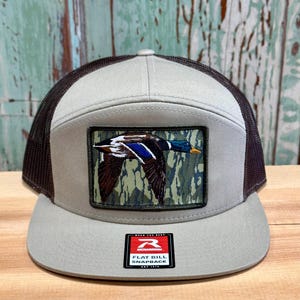 May include: A brown and tan flat bill snapback hat with a camouflage patch featuring a flying mallard duck. The hat has a black mesh back and a Richardson brand label.