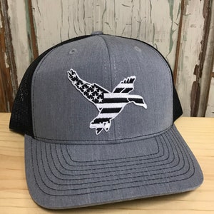 Puede incluir: Gorra de camionero gris con parte trasera de malla negra y visera curva. La gorra presenta un parche bordado de un pato en vuelo, con un diseño que incorpora la bandera estadounidense. La gorra está hecha de tela duradera y tiene un cierre a presión.