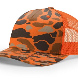 May include: Orange and brown camouflage baseball cap with a mesh back. The cap features a curved brim and a classic trucker hat design. The camouflage pattern includes shades of brown, tan, and orange.