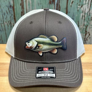 May include: A brown and white trucker hat with a largemouth bass graphic. The hat has a mesh back and an adjustable strap with the Richardson 112 logo.