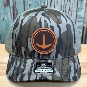 May include: A camouflage trucker hat with a brown leather patch featuring a duck silhouette. The patch has a stitched border and the text "Richardson 112 Original Trucker Adjustable" is on the front of the hat.