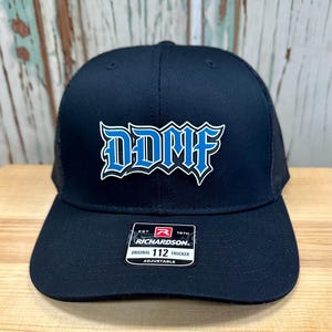 May include: A black trucker hat with a curved brim and mesh back. The front features the blue and white text "DDMF" in a stylized font. A small tag on the brim reads "Richardson Original 112 Trucker Adjustable."