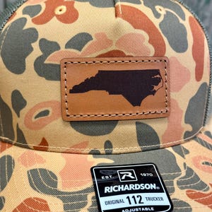 May include: A camouflage trucker hat with a brown leather patch featuring the outline of North Carolina. The hat has a mesh back and a curved brim. The patch is stitched onto the front of the hat. The Richardson brand tag is visible.