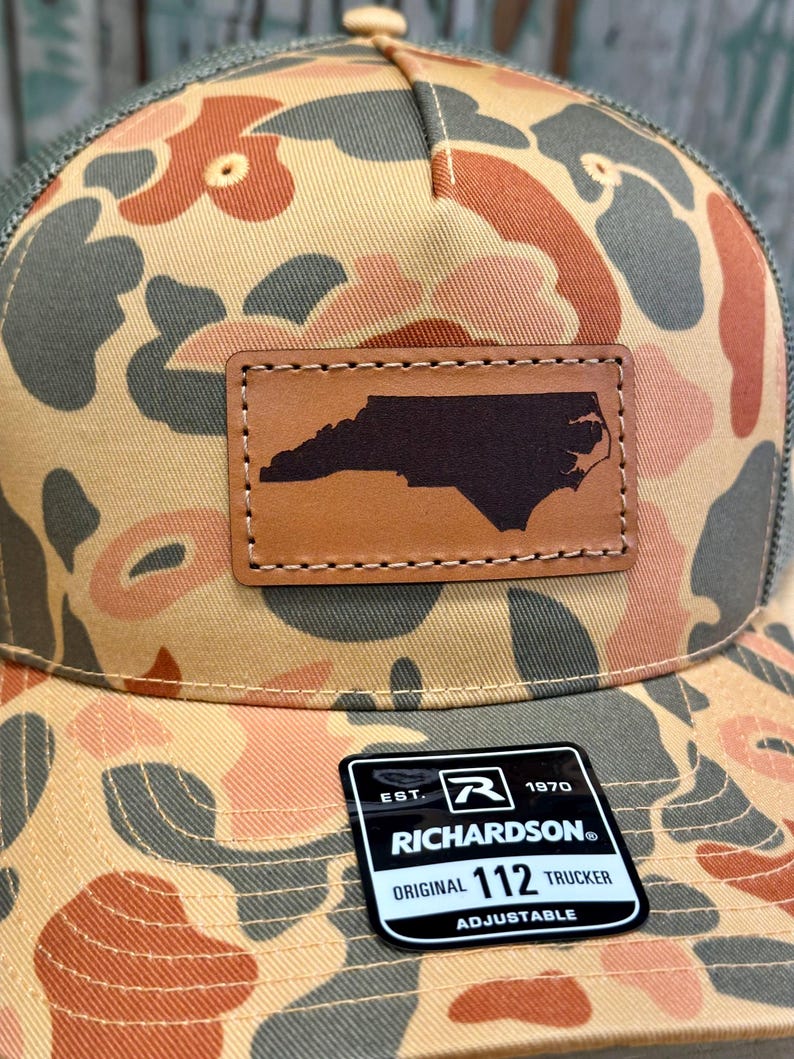 May include: A camouflage trucker hat with a brown leather patch featuring the outline of North Carolina. The hat has a mesh back and a curved brim. The patch is stitched onto the front of the hat. The Richardson brand tag is visible.