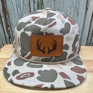 May include: A camouflage-patterned baseball cap with a brown leather patch featuring a deer antler design. The cap has a flat brim and a rope detail around the crown. The color palette includes shades of green, brown, and beige.