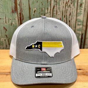 May include: Gray and white trucker hat with a North Carolina state outline patch in black, yellow, and white with the letters "N*C" in the center. The hat has a black label with the text "Richardson" and "Original 112 Trucker Adjustable".