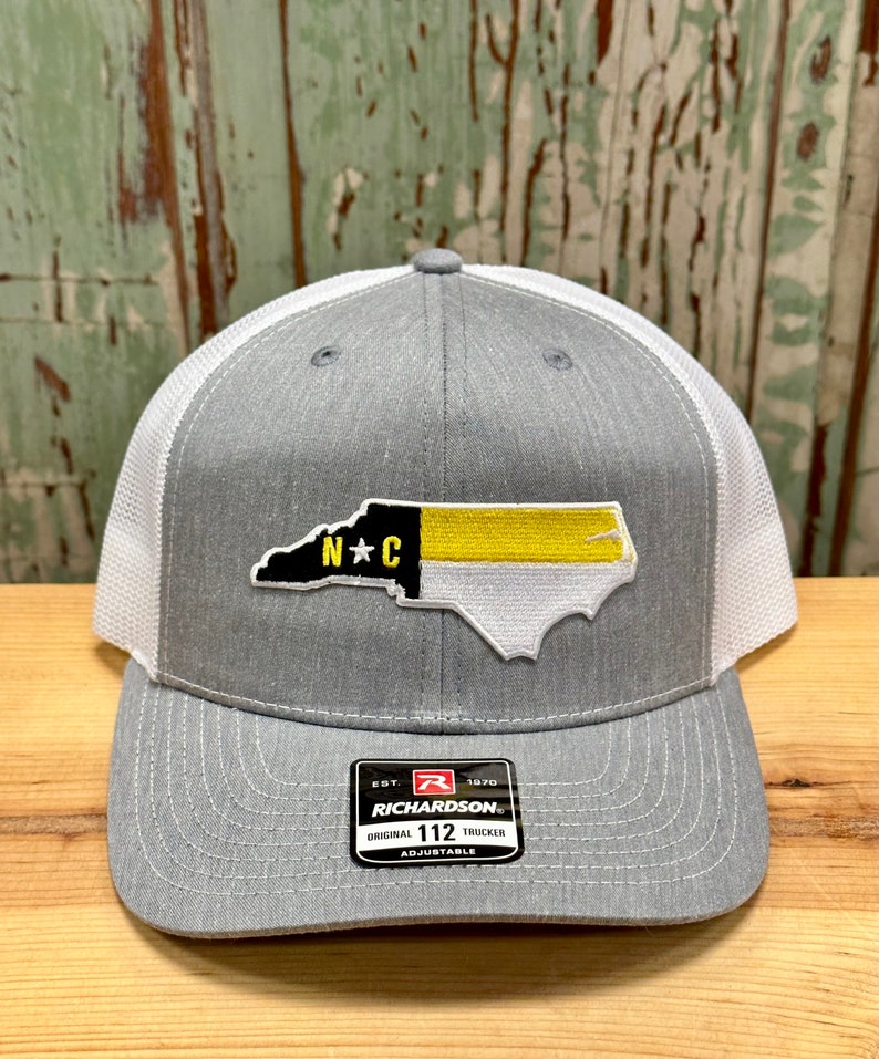 May include: Gray and white trucker hat with a North Carolina state outline patch in black, yellow, and white with the letters "N*C" in the center. The hat has a black label with the text "Richardson" and "Original 112 Trucker Adjustable".
