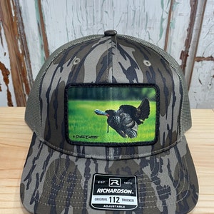 May include: A camouflage trucker hat with a patch featuring a flying wild turkey. The hat is adjustable and has the text "Richardson" and "Original 112 Trucker" on the label.