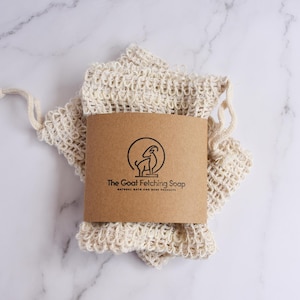 May include: A natural-colored, woven soap bag with a drawstring closure, paired with a brown paper label. The label features a goat illustration and the text "The Goat Fetching Soap". The bag is set against a white marble background.