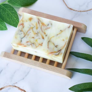 May include: A white bar of soap with yellow and orange swirls sits on a wooden soap dish. The soap dish is made of light brown wood and has a slatted design. Green leaves are visible in the background.