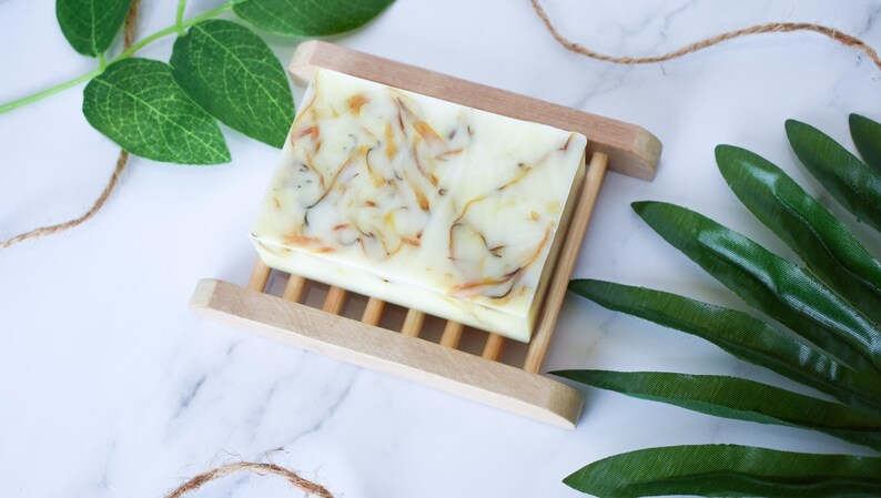 May include: A white bar of soap with yellow and orange swirls sits on a wooden soap dish. The soap dish is made of light brown wood and has a slatted design. Green leaves are visible in the background.
