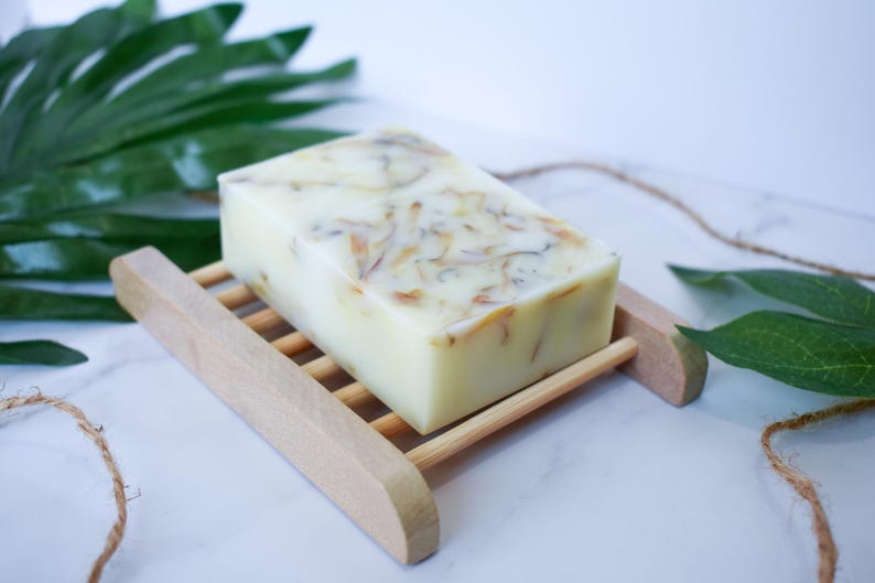 May include: A white bar of soap with dried flowers and herbs embedded in it. The soap is sitting on a wooden soap dish.