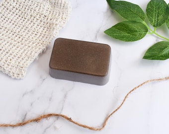 May include: A rectangular brown soap bar sits next to a beige mesh washcloth and a green leafy sprig on a white marble surface. A length of brown twine is also present. The soap appears to be handmade and is the focal point of the image.