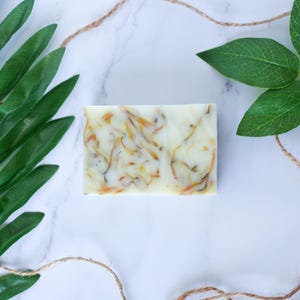 May include: A white bar of soap with swirls of yellow and green. The soap is on a white marble surface with green leaves on either side.