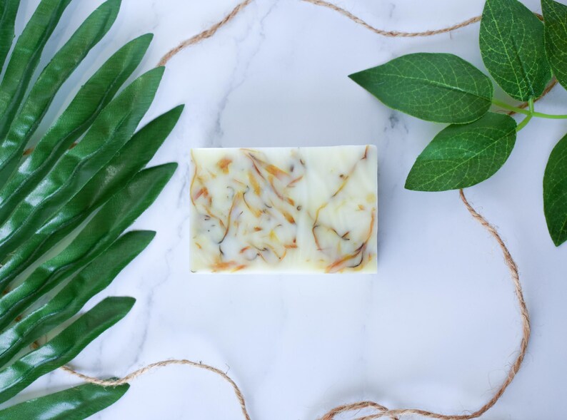 May include: A white bar of soap with swirls of yellow and green. The soap is on a white marble surface with green leaves on either side.