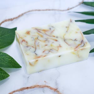 May include: A white bar of soap with swirls of yellow and orange. The soap is on a white surface with green leaves.