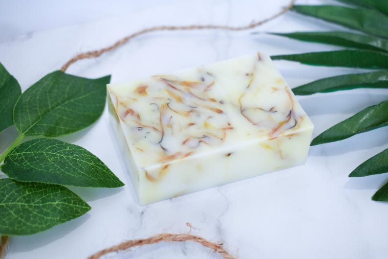 May include: A white bar of soap with swirls of yellow and orange. The soap is on a white surface with green leaves.