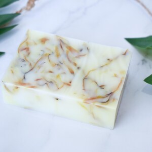 May include: A rectangular bar of soap with a white base and swirls of brown and yellow throughout. The soap is on a white marble surface with green leaves in the background.