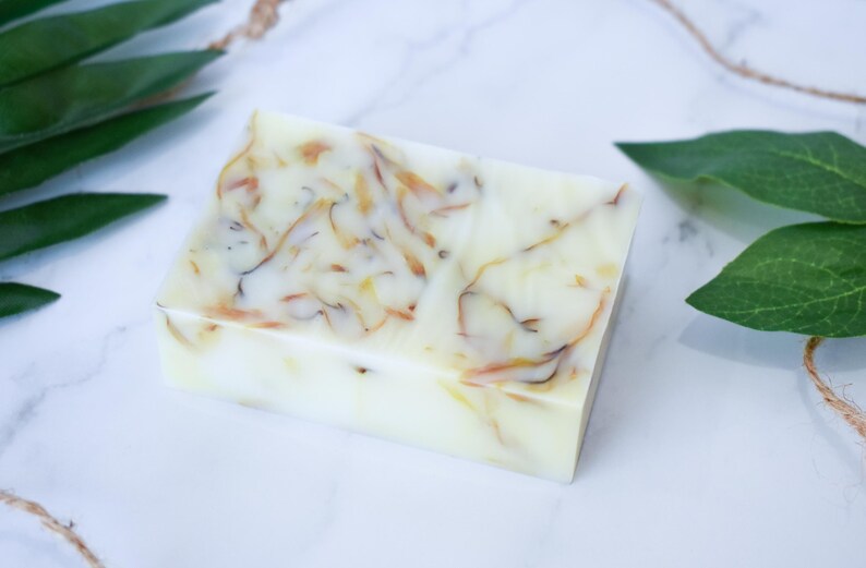 May include: A rectangular bar of soap with a white base and swirls of brown and yellow throughout. The soap is on a white marble surface with green leaves in the background.