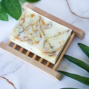 May include: A white bar of soap with dried flowers and herbs inside, resting on a wooden soap dish. The soap dish is made of light brown wood and has a slatted design. The soap is surrounded by green leaves.