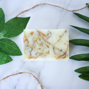 May include: A white bar of soap with swirls of yellow and orange. The soap is on a white marble surface with green leaves.