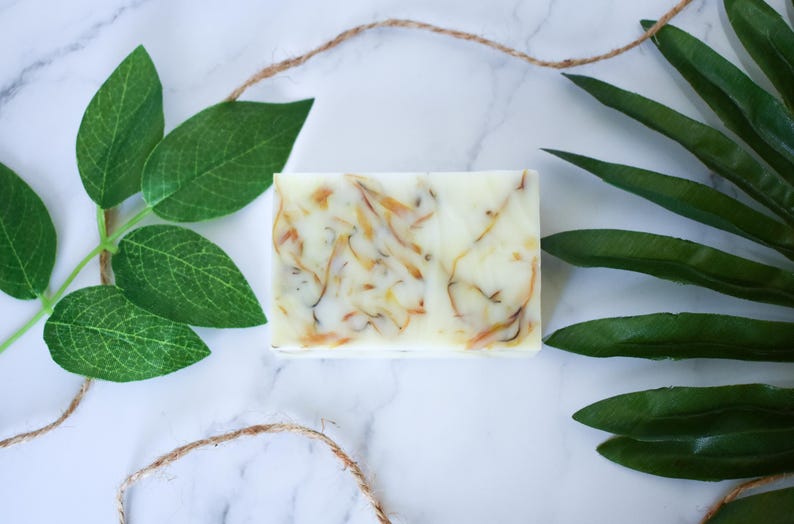 May include: A white bar of soap with swirls of yellow and orange. The soap is on a white marble surface with green leaves.