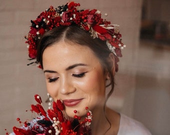 May include: A bride wearing a white dress holds a bouquet of red and pink dried flowers. The bouquet is made of red berries, pink roses, and other dried flowers. The bride is also wearing a red flower crown.
