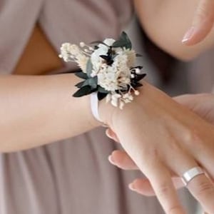 May include: A white and green dried flower wrist corsage with a white satin ribbon.